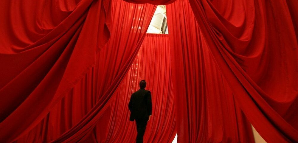 Un homme se tient devant une installation artistique composée de grands rideaux rouges rappelant ceux d’un théâtre.