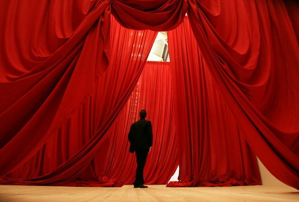 Un homme se tient devant une installation artistique composée de grands rideaux rouges rappelant ceux d’un théâtre.