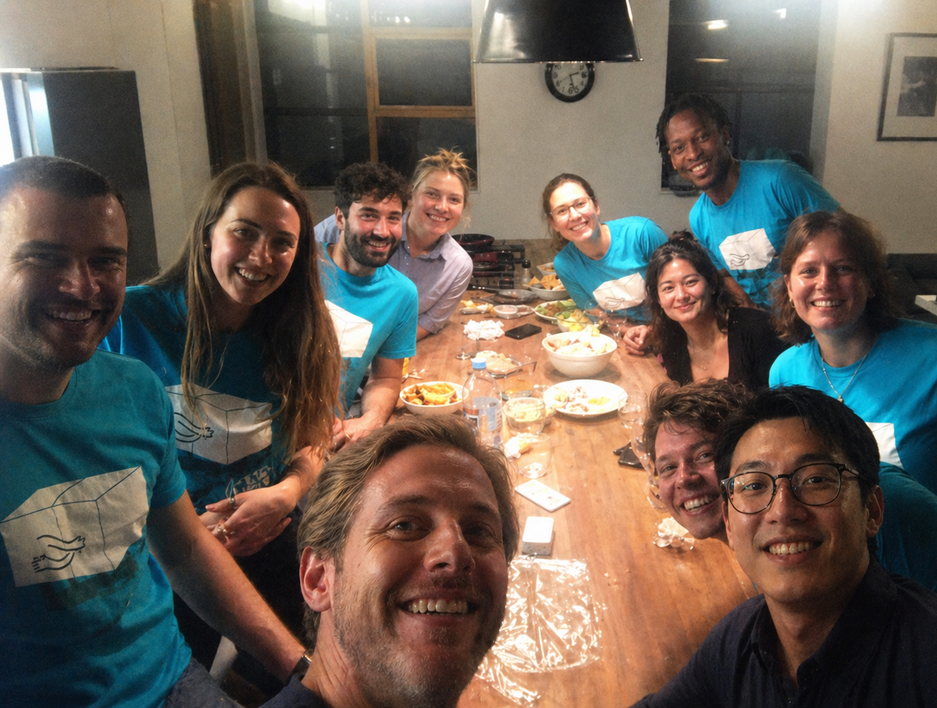 L'équipe de locationWorkshop souriante autour d'une table de dîner, portant tous des t-shirts turquoise assortis avec le logo Tetrix représentant deux cubes qui s'embrassent.